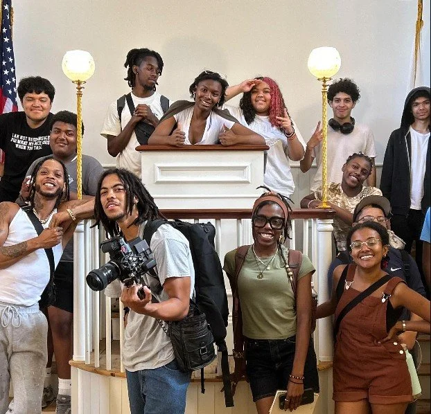 Group of young people smiling and posing inside a building, some standing and some sitting, with a white panel and two tall lamps in the background.
