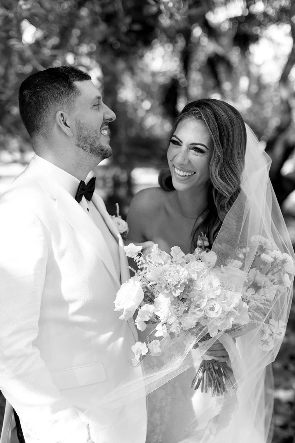 A black and white photo of a bride and groom smiling and looking at each other outdoors, the bride holding a bouquet of flowers and wearing a veil.