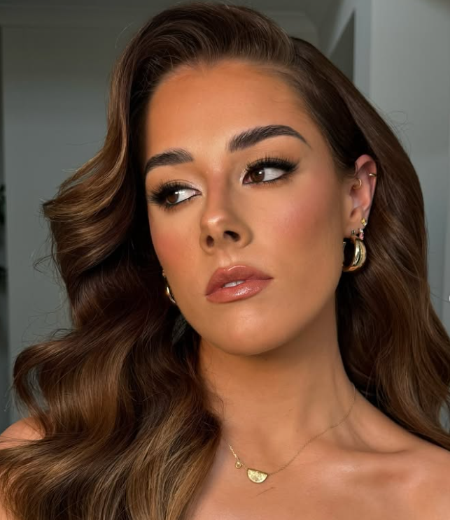 A woman with long, wavy brown hair and light makeup, wearing gold hoop earrings and a gold necklace, looking to the side in an indoor setting.