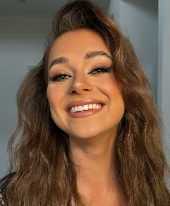 Close-up of a smiling woman with wavy brown hair, makeup, and a shiny lipstick, indoors.