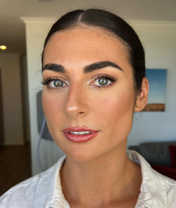 Close-up portrait of a woman with blue eyes, dark hair pulled back, wearing a white shirt, in a well-lit room with a blurred background.