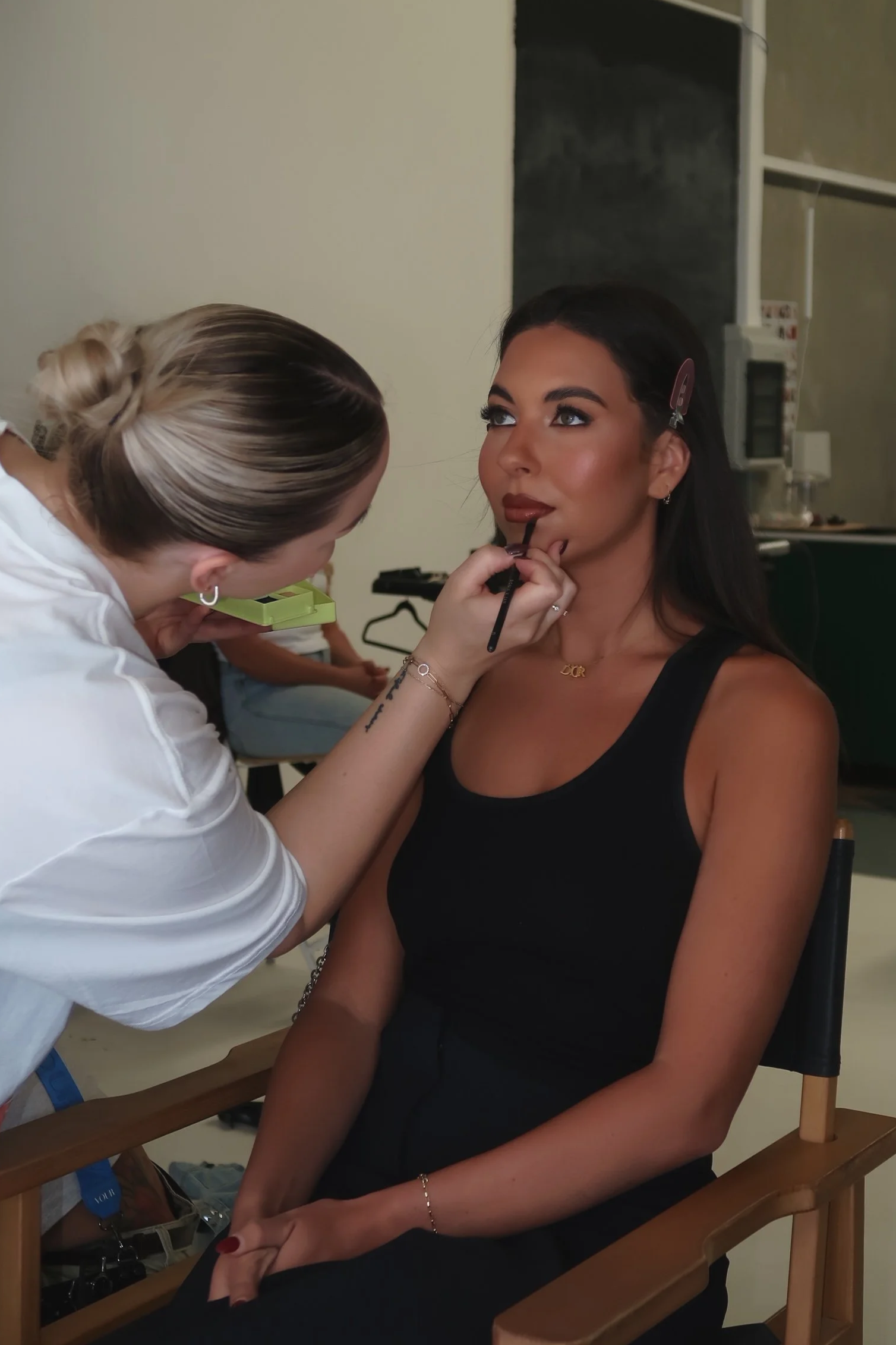 A woman with dark hair sitting in a chair getting her makeup done by a makeup artist. The makeup artist is focused on applying lipstick, with her head bent close to the woman's lips. The woman is wearing a black sleeveless top and has a hair clip in her hair.