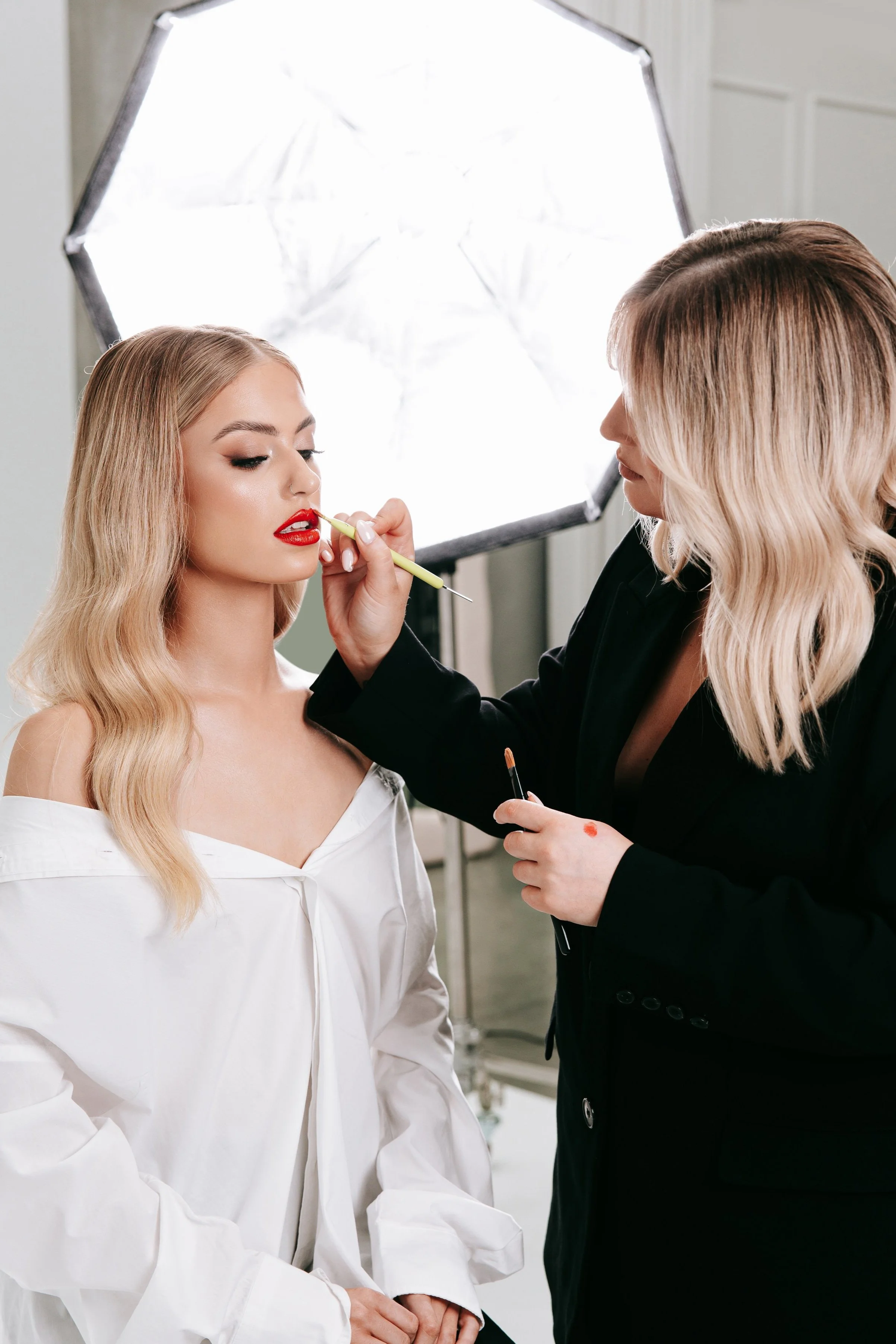 A woman with blonde hair, wearing a white shirt, is getting her makeup done by a makeup artist. The makeup artist is applying red lipstick. A photography light is visible in the background.