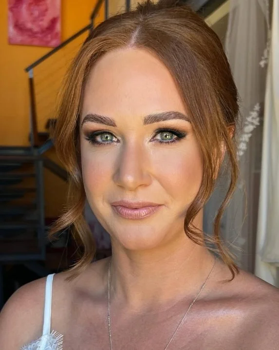 Close-up of a woman with fair skin, red hair, and blue eyes, smiling indoors with warm lighting and modern decor in the background.