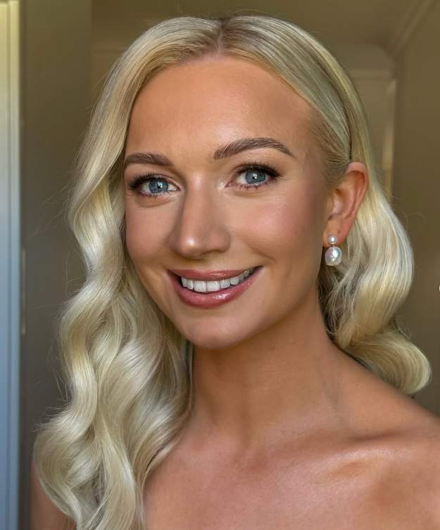 A woman with long blonde wavy hair, blue eyes, and fair skin, smiling, wearing pearl earrings, in an indoor setting.