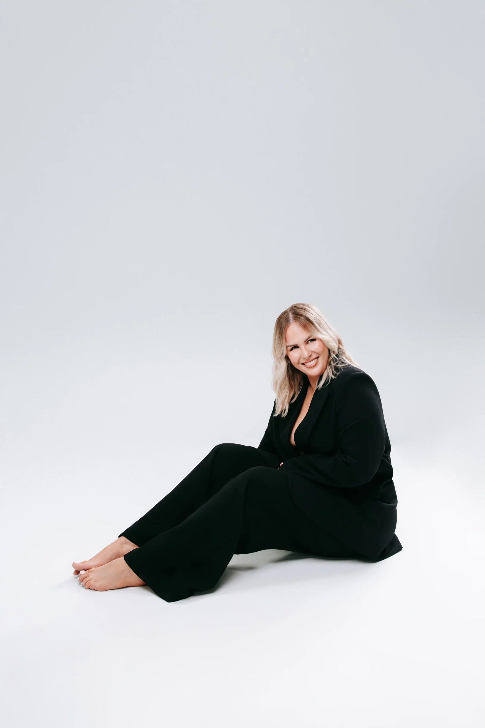 A woman with blonde hair, sitting on the floor, smiling, wearing a black blazer and pants, barefoot, against a plain light gray background.