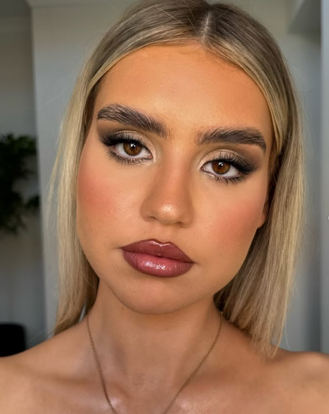 Close-up of a woman with blonde hair, brown eyes, full lips with gloss, and makeup emphasizing her eyes and eyebrows.
