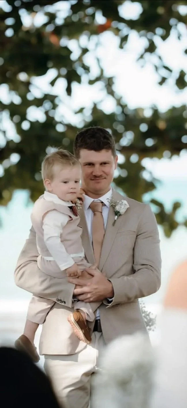 A man holding a young boy at an outdoor wedding ceremony with trees in the background.