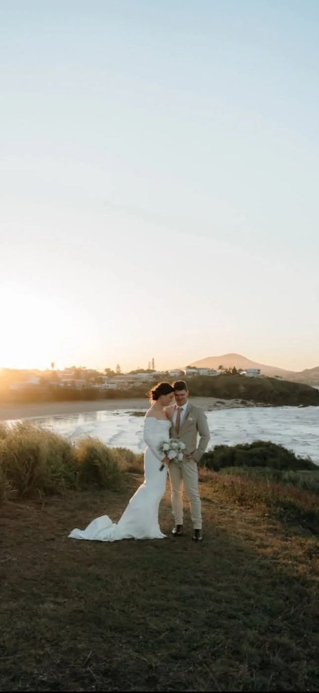 A bride and groom standing close together on a grassy coastal area during sunset. The bride is in a white wedding dress holding a bouquet, and the groom is in a light-colored suit. The background features the ocean, coastline, and a distant town or h