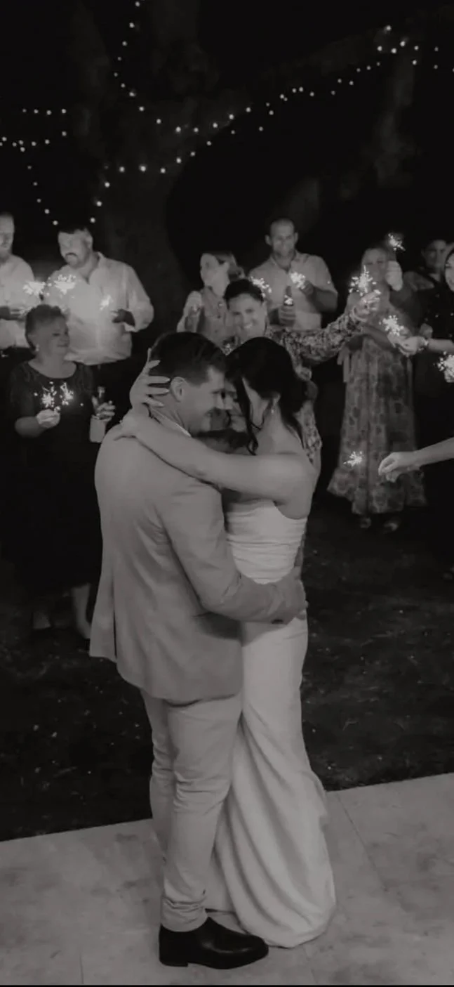 A couple dancing closely at a nighttime celebration with friends holding sparklers around them, under string lights hanging from a large tree.