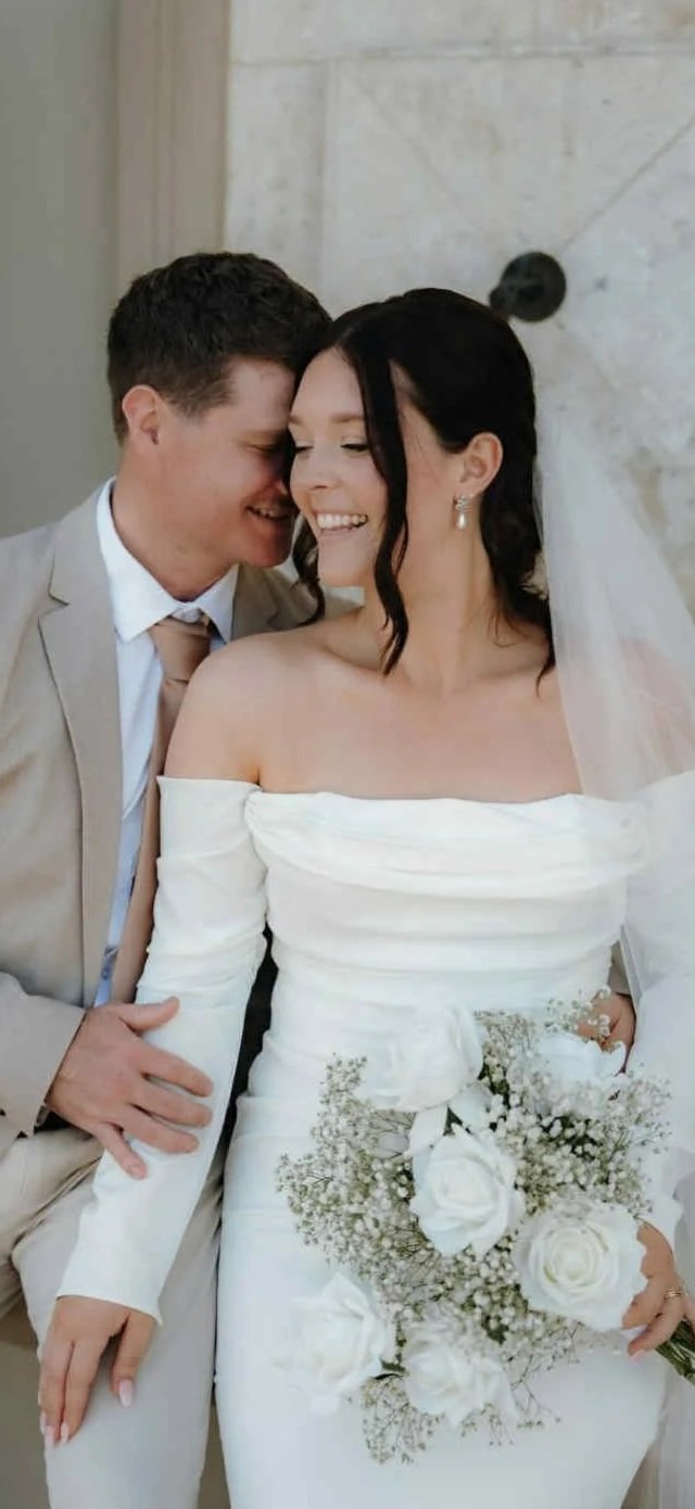 A bride and groom sharing a joyful moment, with the groom whispering to the bride who is smiling and holding a floral bouquet, in a wedding setting.