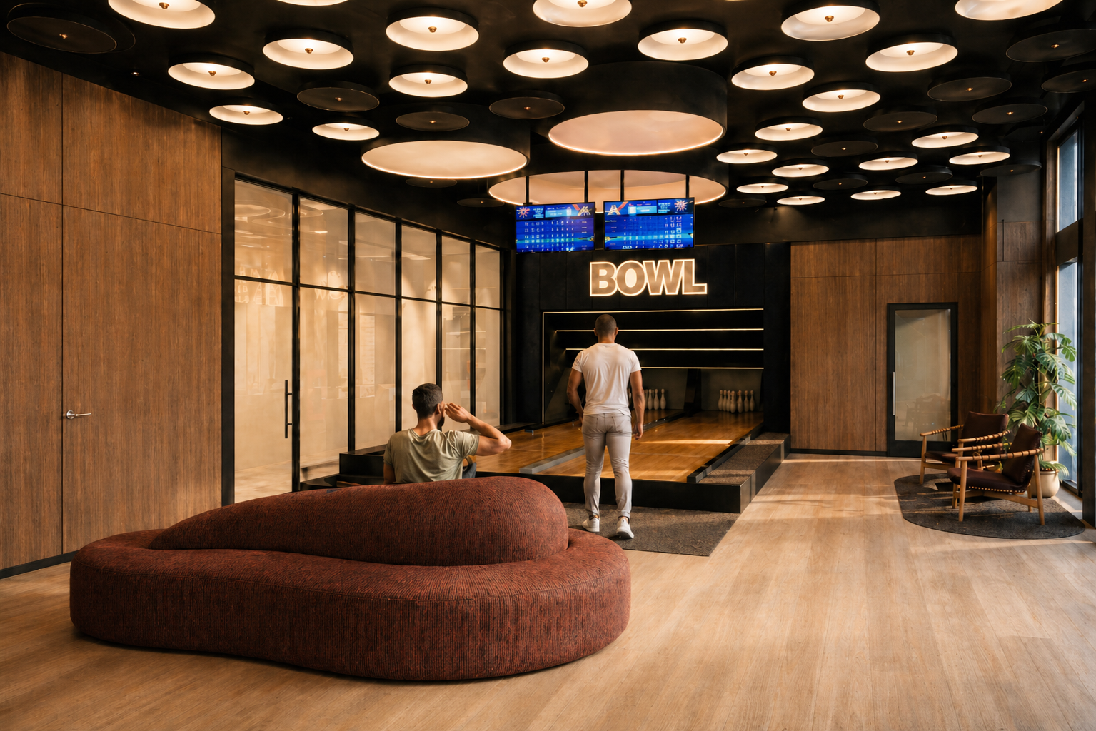 Interior of a modern bowling alley with wooden walls and floors, large circular ceiling lights, a digital scoreboard, and a large illuminated sign that says 'BOWL.' There are two men in the scene, one sitting on a curved red sofa and another standing near the bowling lane, which has pins set up at the end.