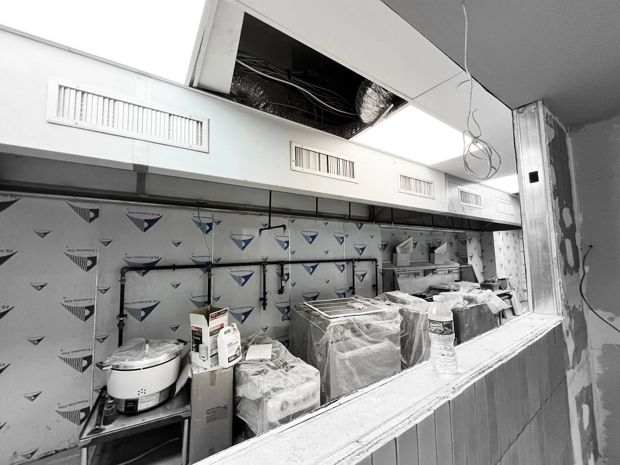 Construction site with exposed ceiling, ventilation ducts, and electrical wiring. Tools and supplies covered with plastic sheets on a countertop.