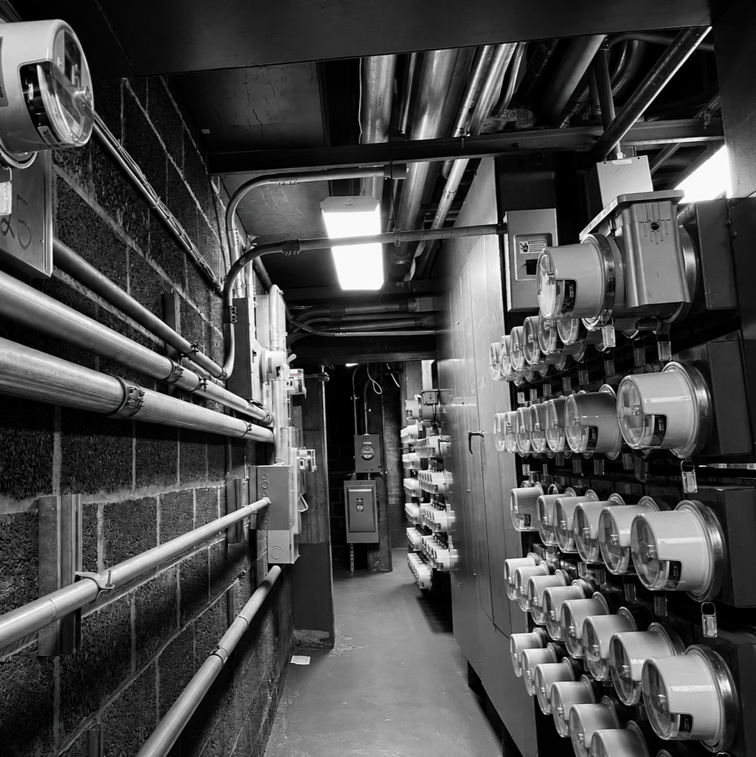 A black and white photo of an industrial electrical and plumbing utility room with pipes, control panels, and electrical meters mounted on the wall.