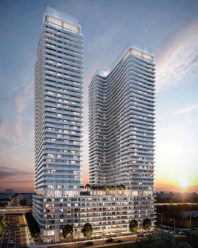 Two modern high-rise buildings with glass facades and exterior balconies, set against a sunset sky with a cityscape and street visible at the base.