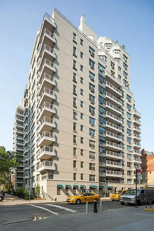 Tall, modern white apartment building with multiple balconies on each floor, located on a city street with yellow taxis and cars parked in front.