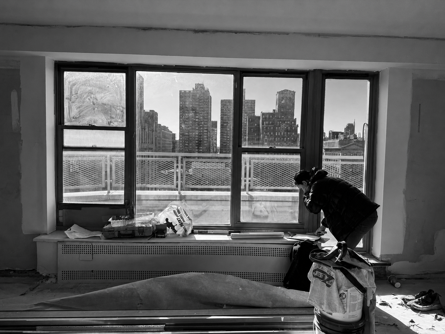 A person is working on a construction or renovation project inside a room with a large window overlooking a cityscape with tall buildings. The room appears unfinished with construction tools, materials, and debris scattered around.