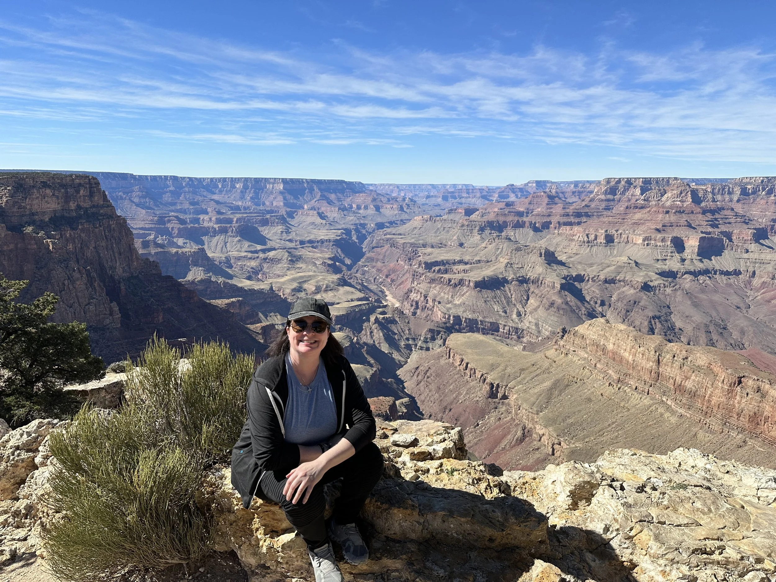 A woman wearing a black hat, sunglasses, a gray t-shirt, a black jacket, and black pants, sitting on rocks at the edge of the Grand Canyon with a smile, surrounded by desert vegetation, under a blue sky with wispy clouds.