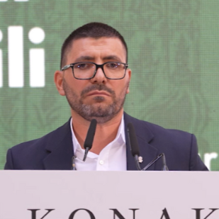 A man wearing a hat and glasses sits at a table with a microphone, water bottle, and plastic cup in front of him.