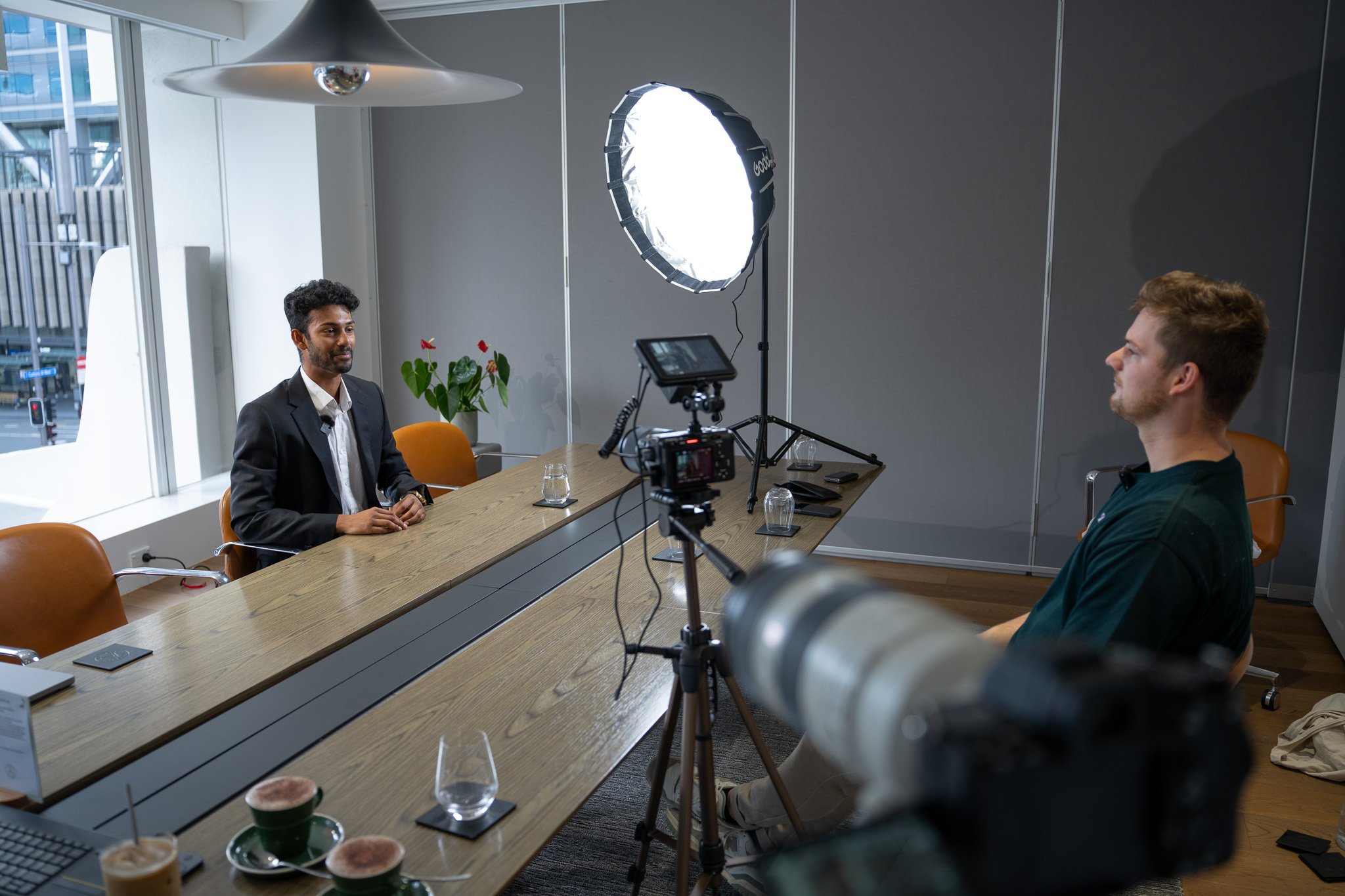 A man wearing a suit sitting at a conference table being recorded during an interview or video shoot, with professional lighting and camera equipment set up in a modern office setting.