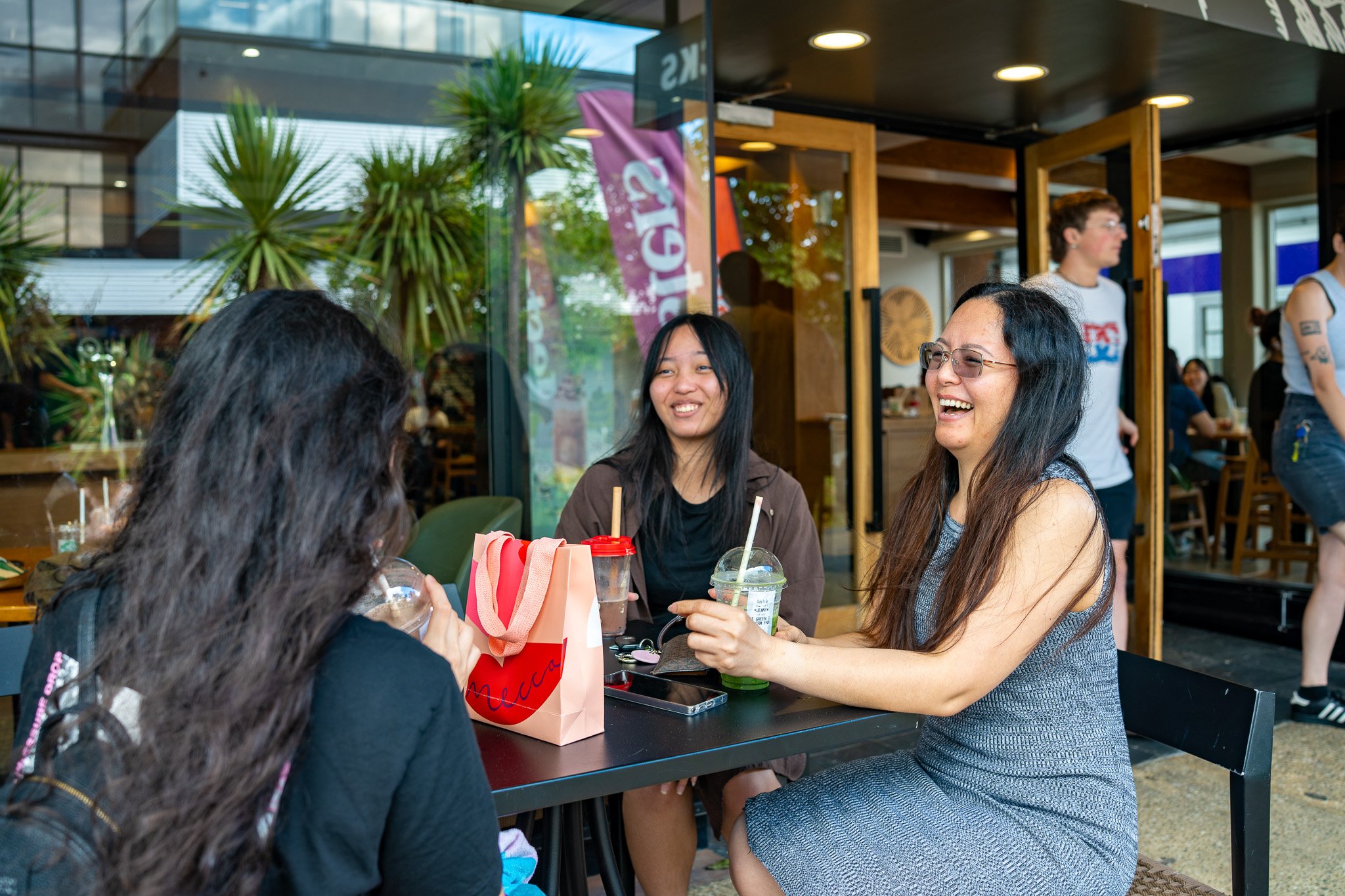 Three women sitting at a table in a café, smiling and chatting, with two of them holding drinks, and a shopping bag on the table. In the background, people are walking and sitting inside the restaurant.
