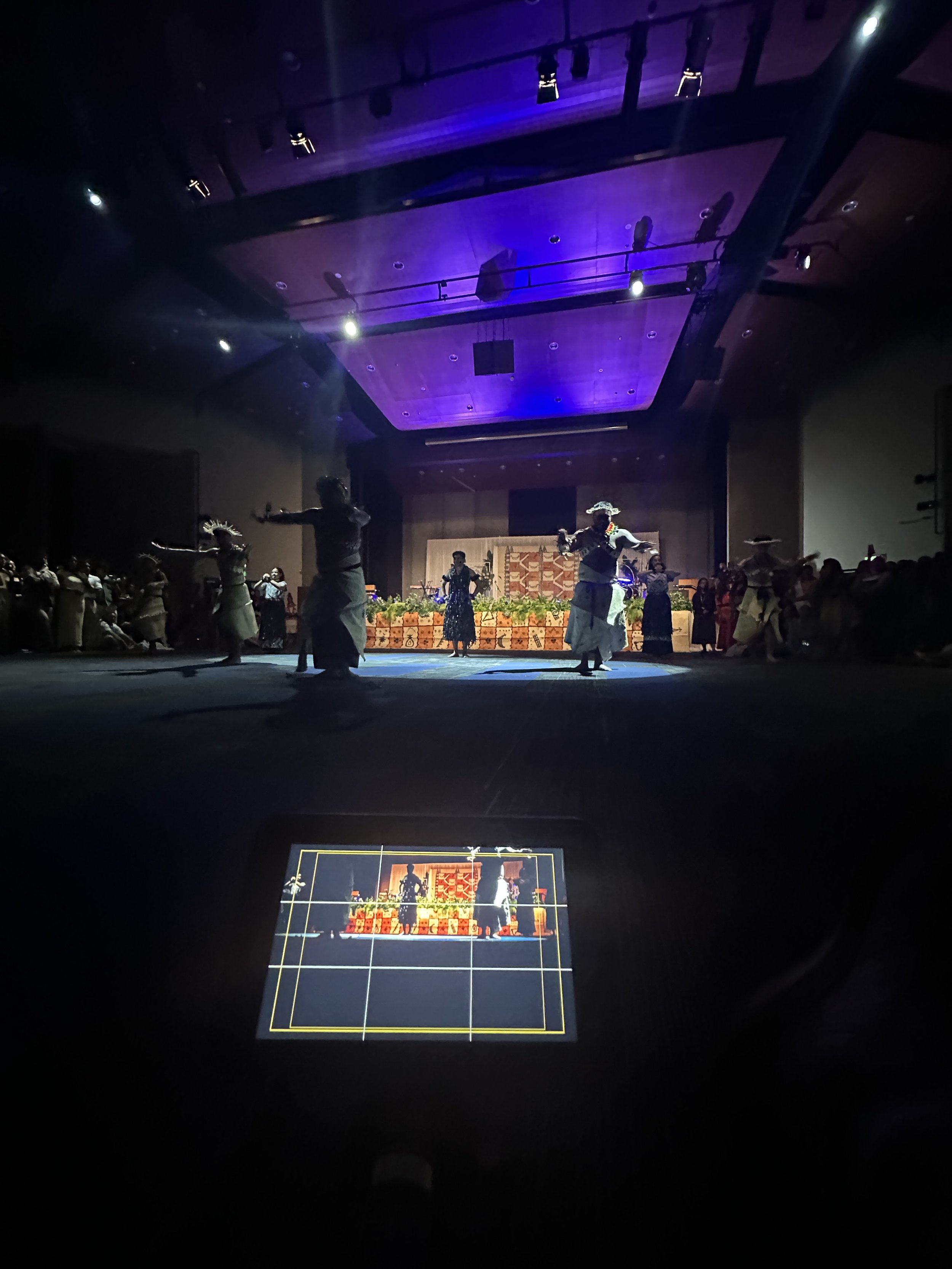 A group of dancers performing on an indoor stage with a purple-lit ceiling and a decorated backdrop. Audience members watch from the sides, with a camera or device screen displaying a grid overlay and a live view of the dancers.