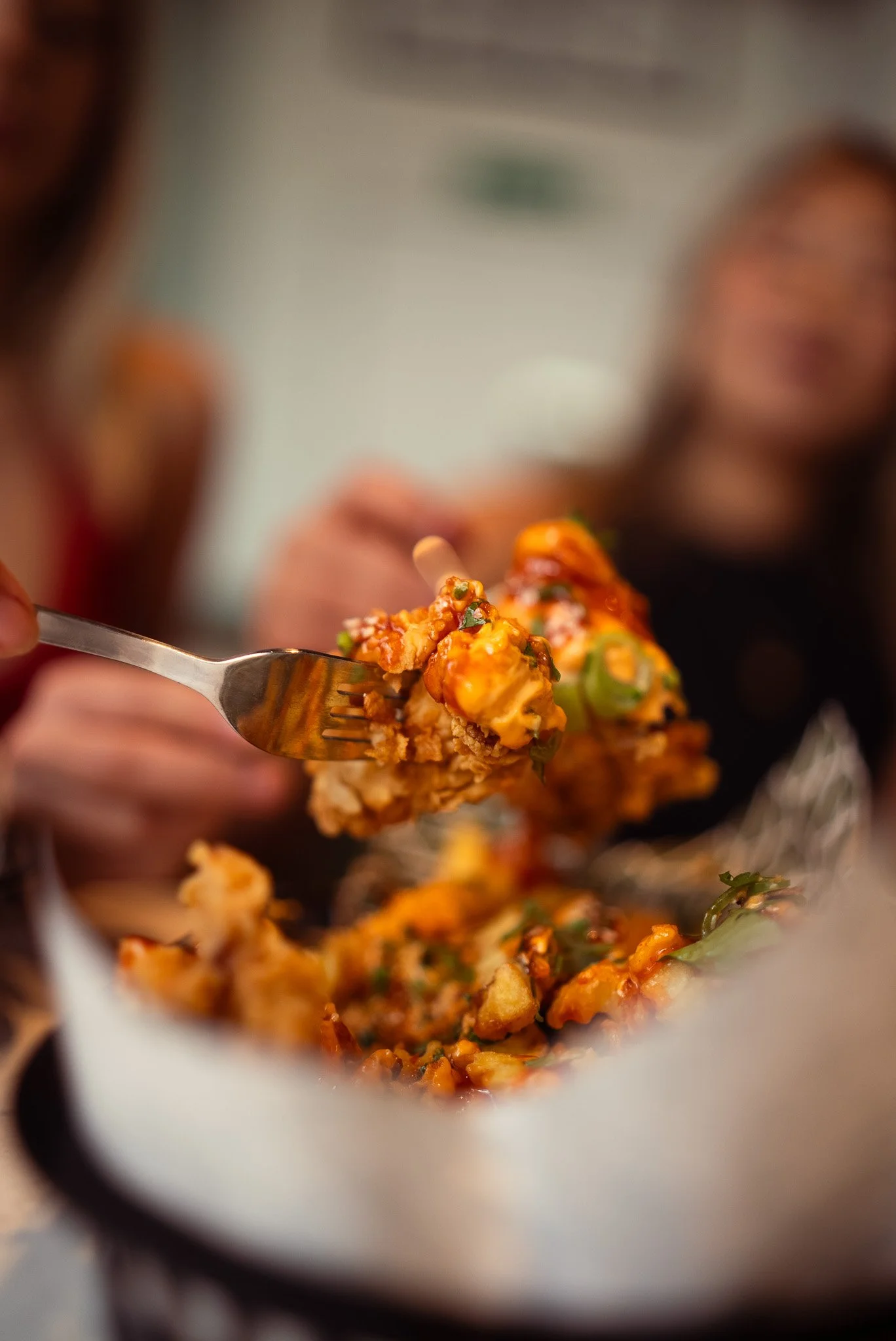 A fork with a piece of fried chicken topped with sauce and garnished with green onions, held over a paper takeout container filled with more chicken pieces in sauce.