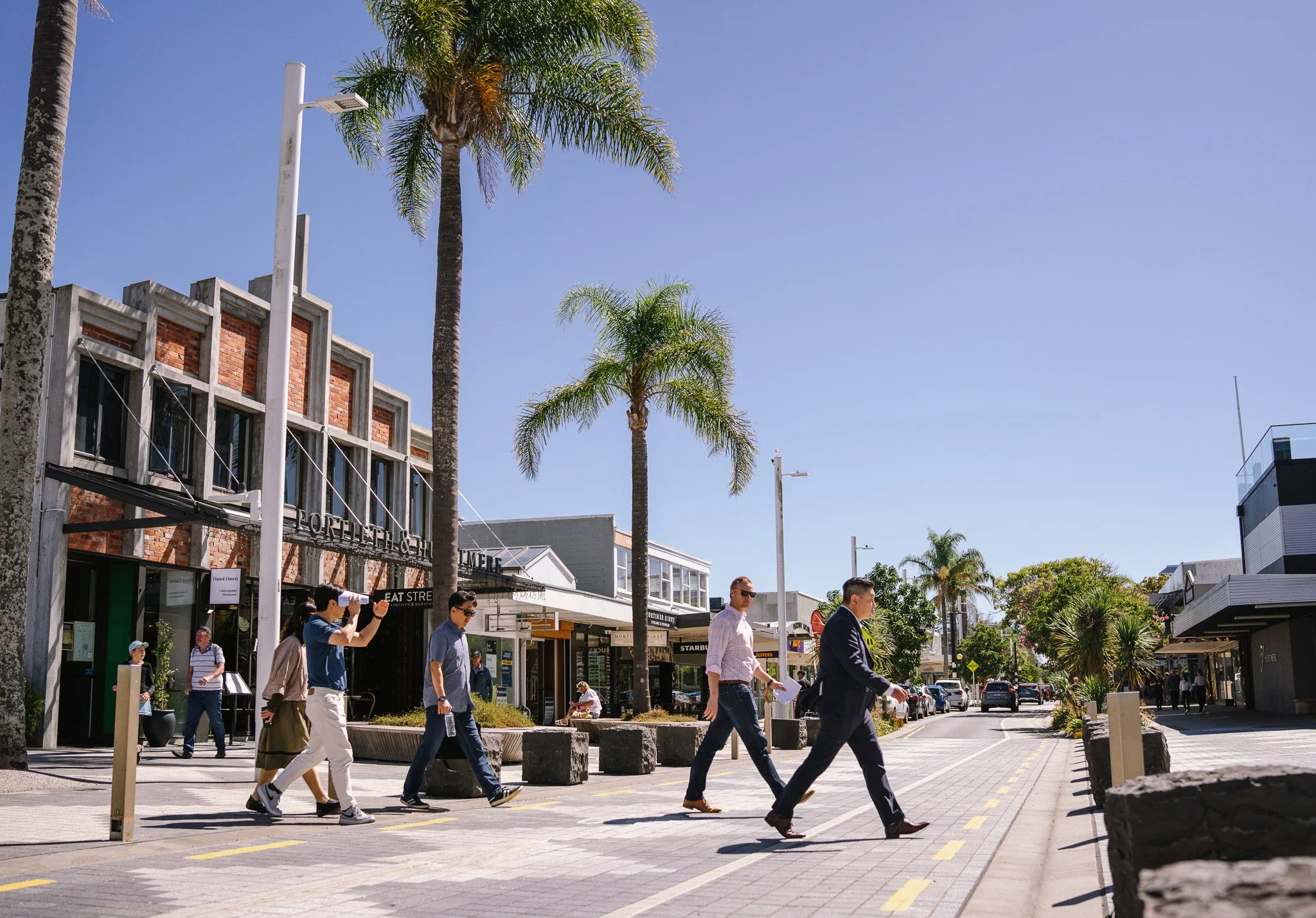 People walking across a crosswalk on a sunny day in a shopping district with palm trees and storefronts.