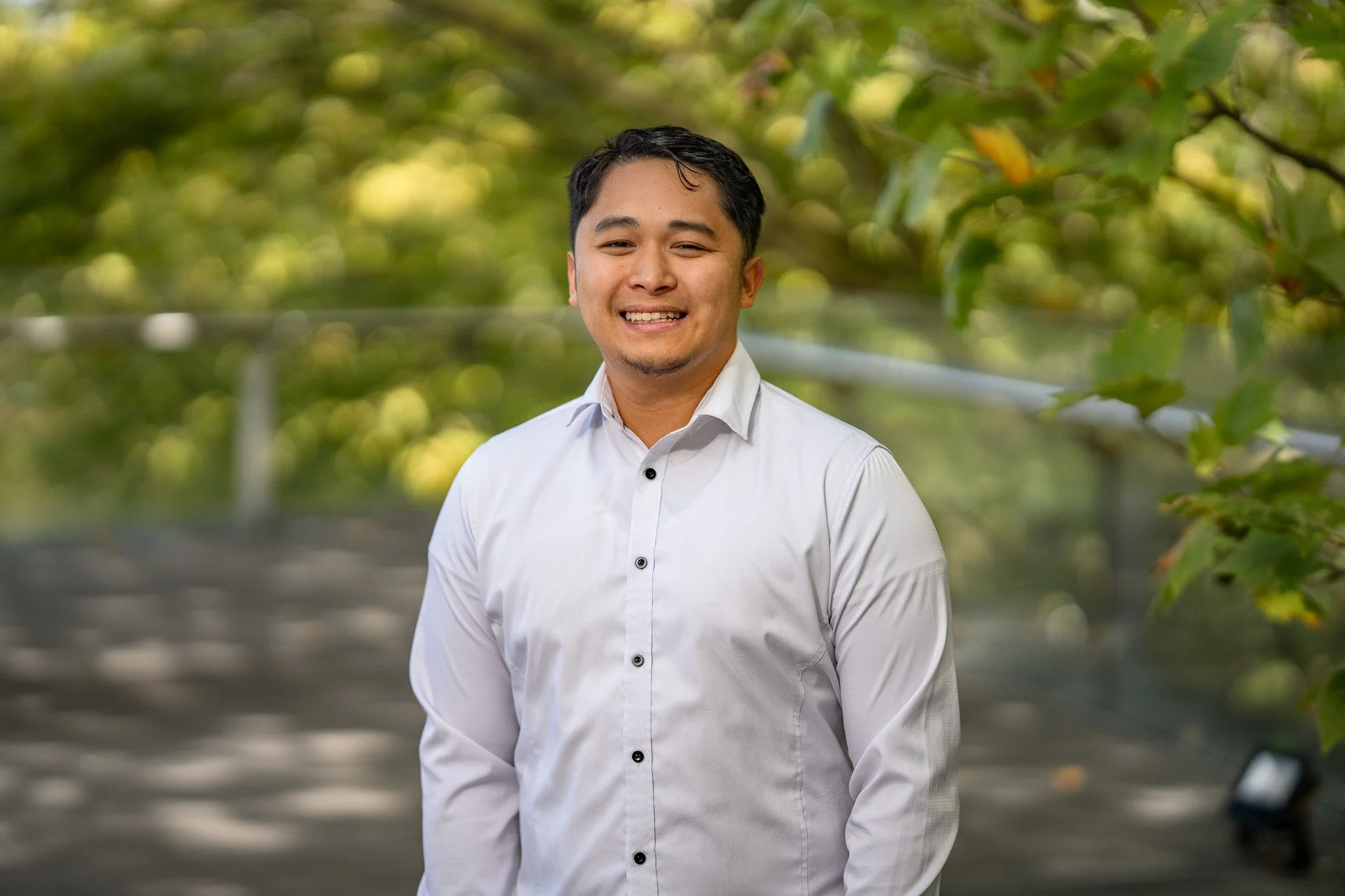 A man in a white button-up shirt smiling outdoors with green foliage in the background.