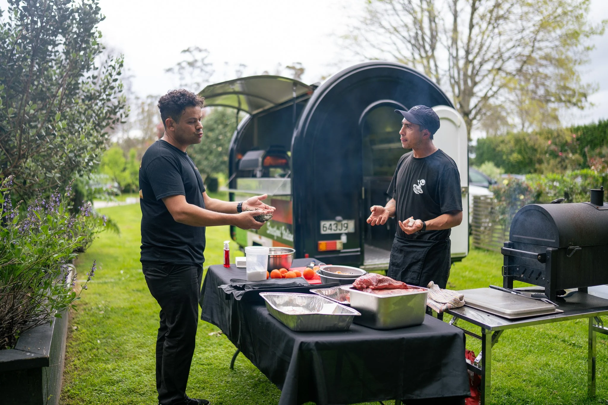 Two men standing next to each other outdoors near a table with food, a barbecue grill, and a small trailer in the background. One man is handing over some food, while the other is gesturing.