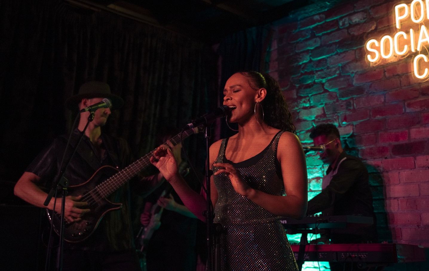 A woman singing into a microphone with eyes closed, wearing a sparkly dress and large hoop earrings, accompanied by two musicians playing guitar and keyboard, on a stage with a brick wall and neon sign in the background.