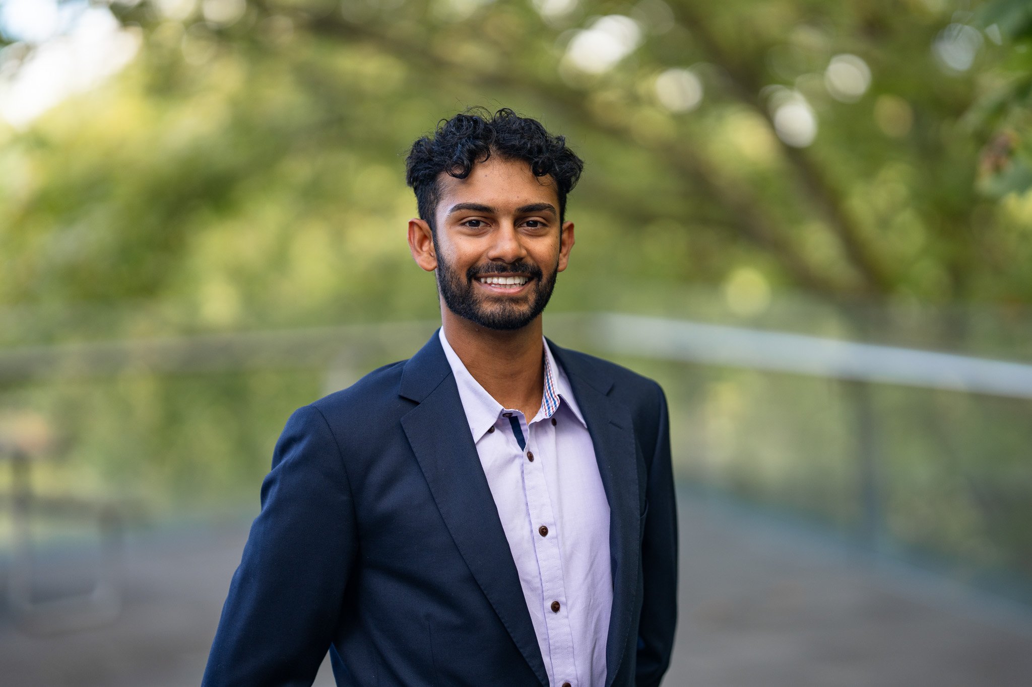 A smiling man in a navy suit standing outdoors with blurred green trees in the background.