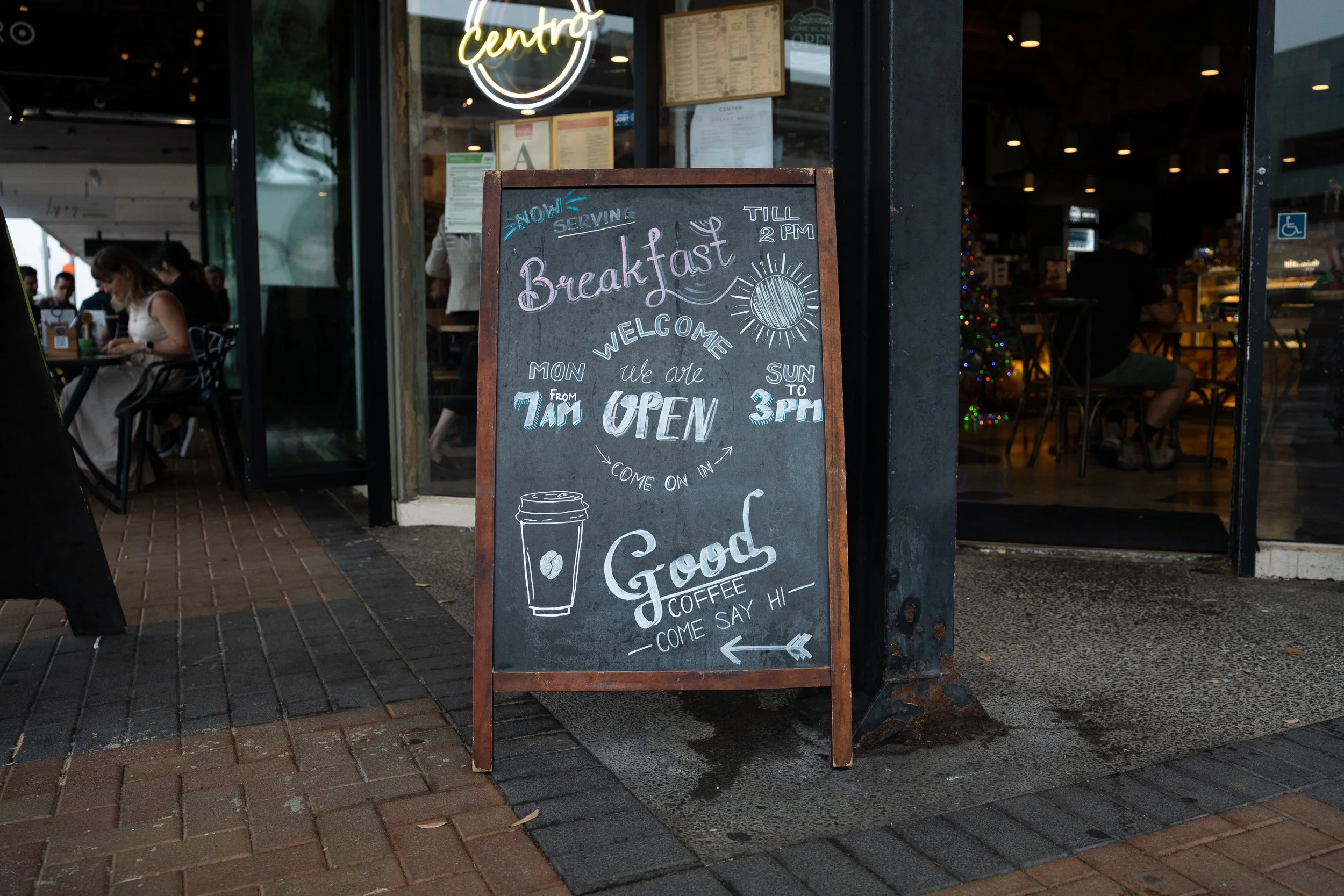 Chalkboard sign outside a cafe advertising breakfast hours, opening from 7 am Monday to Sunday at 3 pm, with drawings of a coffee cup and an arrow.