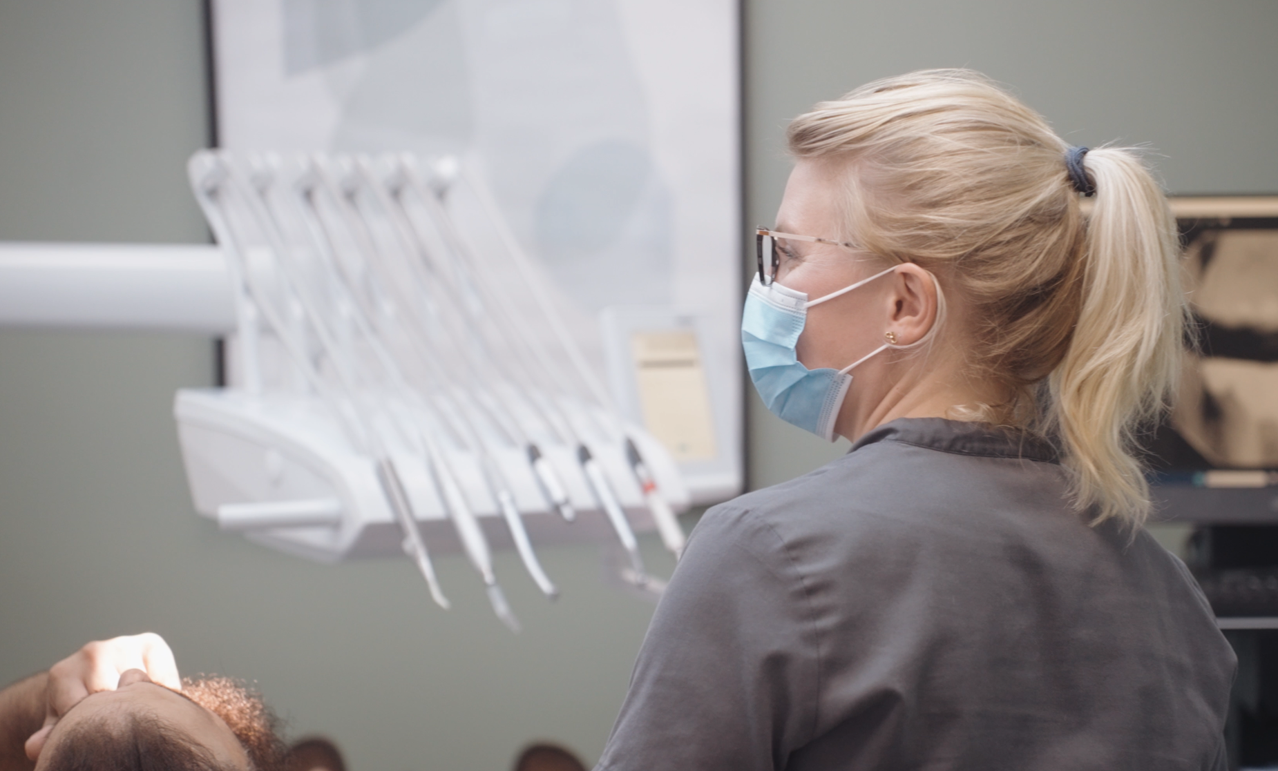 A female dental professional wearing glasses and a face mask examines a patient in a dental clinic.