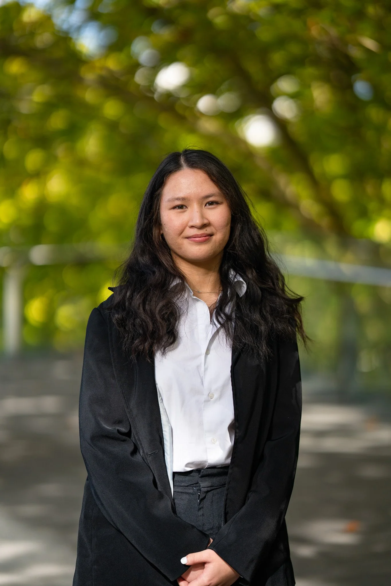 A young woman with long dark wavy hair wearing a black jacket and white shirt, standing outdoors with a blurred green foliage background.