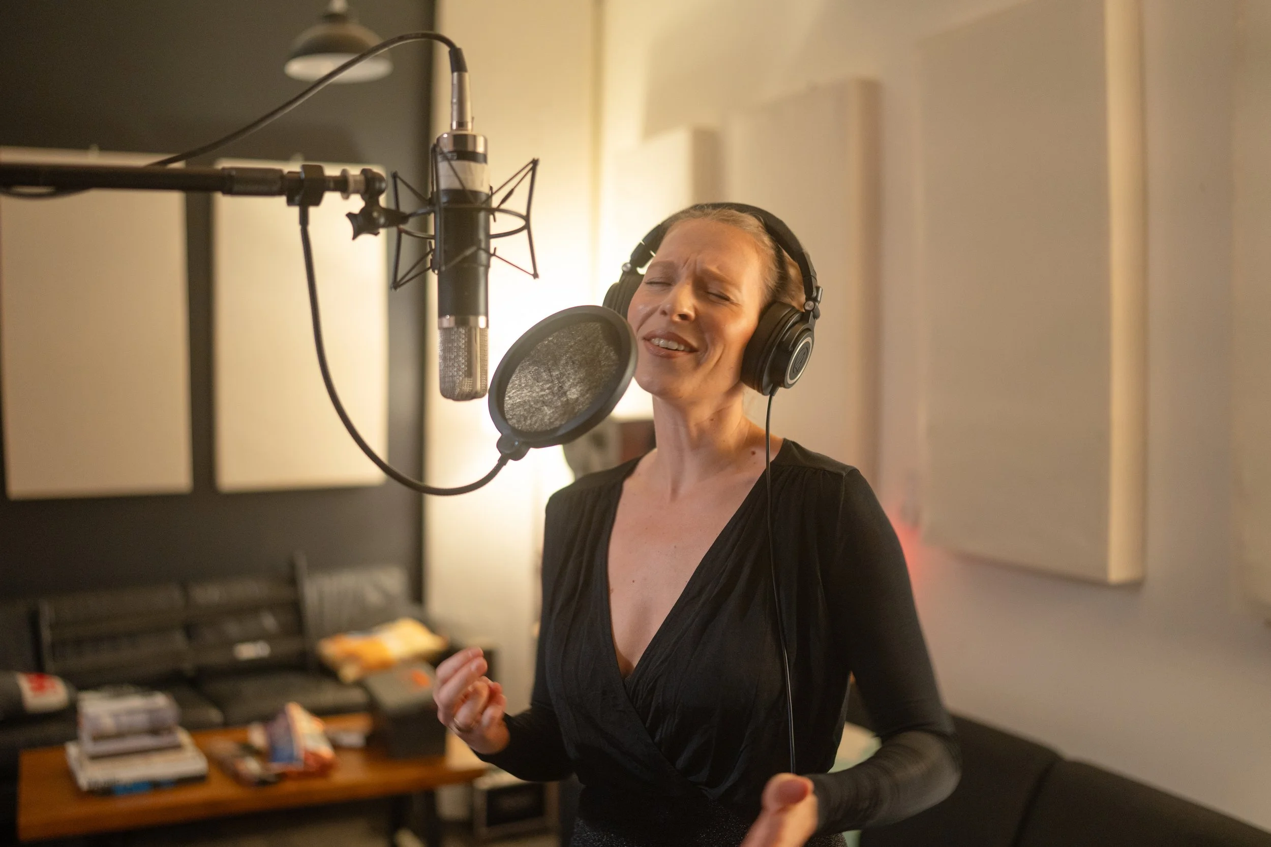 A woman singing into a microphone in a recording studio, wearing headphones, with her eyes closed and an emotional expression.