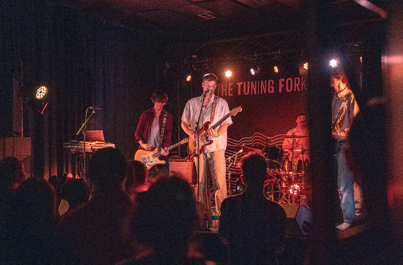 Band performing live on stage at a music venue with the sign 'The Tuning Fork' behind them, featuring four band members playing instruments including guitars and drums, with audience visible in the foreground.
