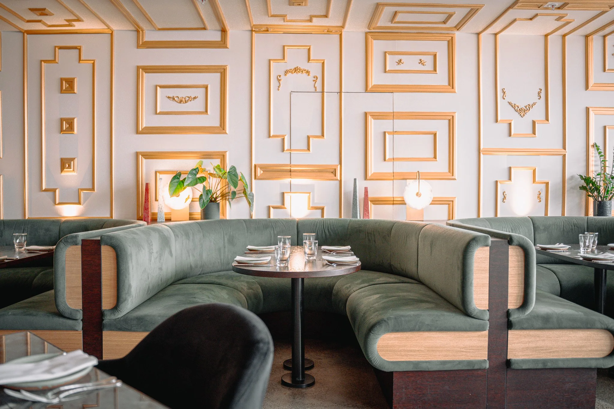 Indoor dining area with green booth seating and tables set with plates, glasses, and silverware. The background features a white wall with gold decorative molding and potted plants.