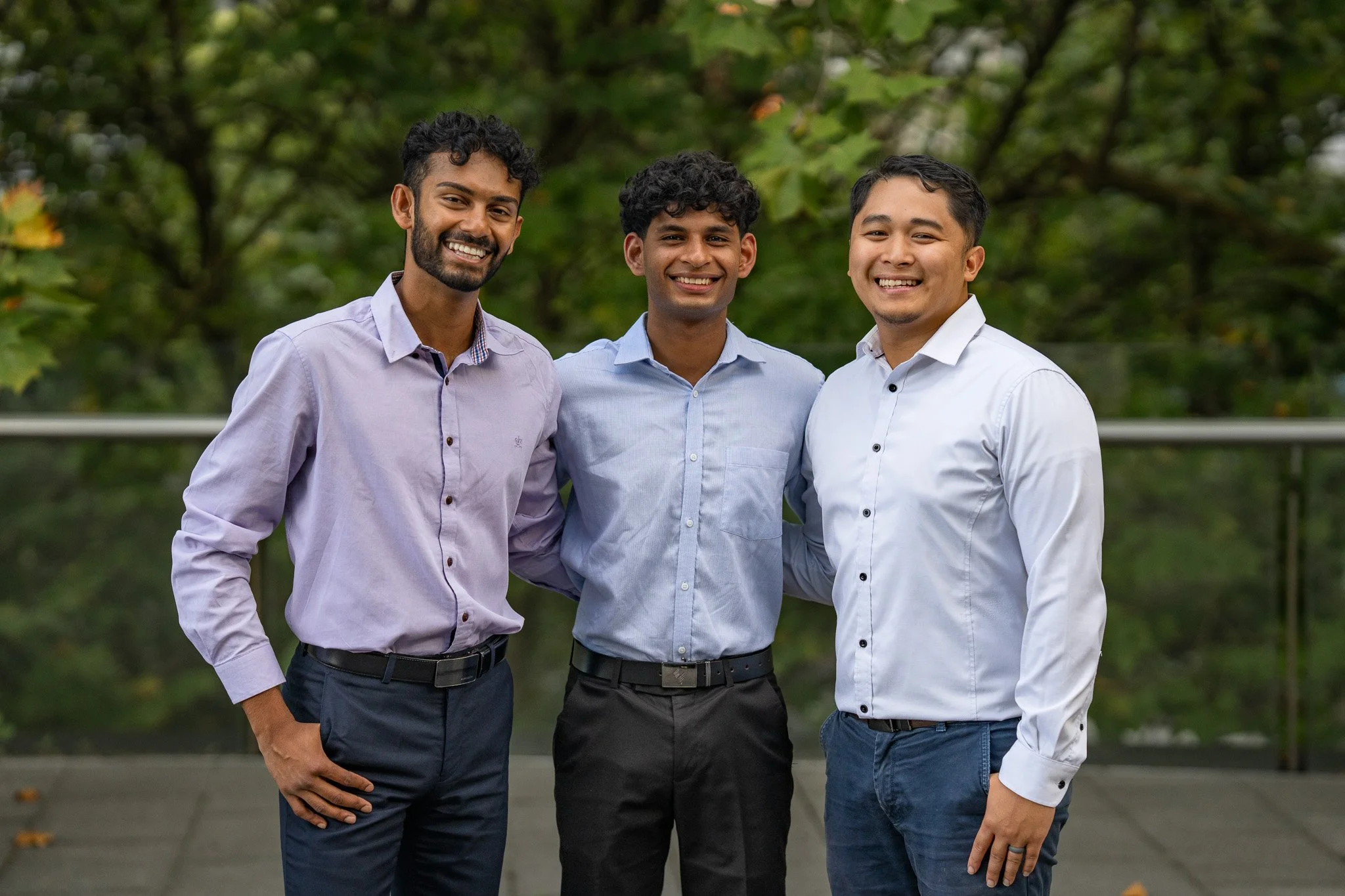 Three young men dressed in business casual attire standing outdoors smiling at the camera
