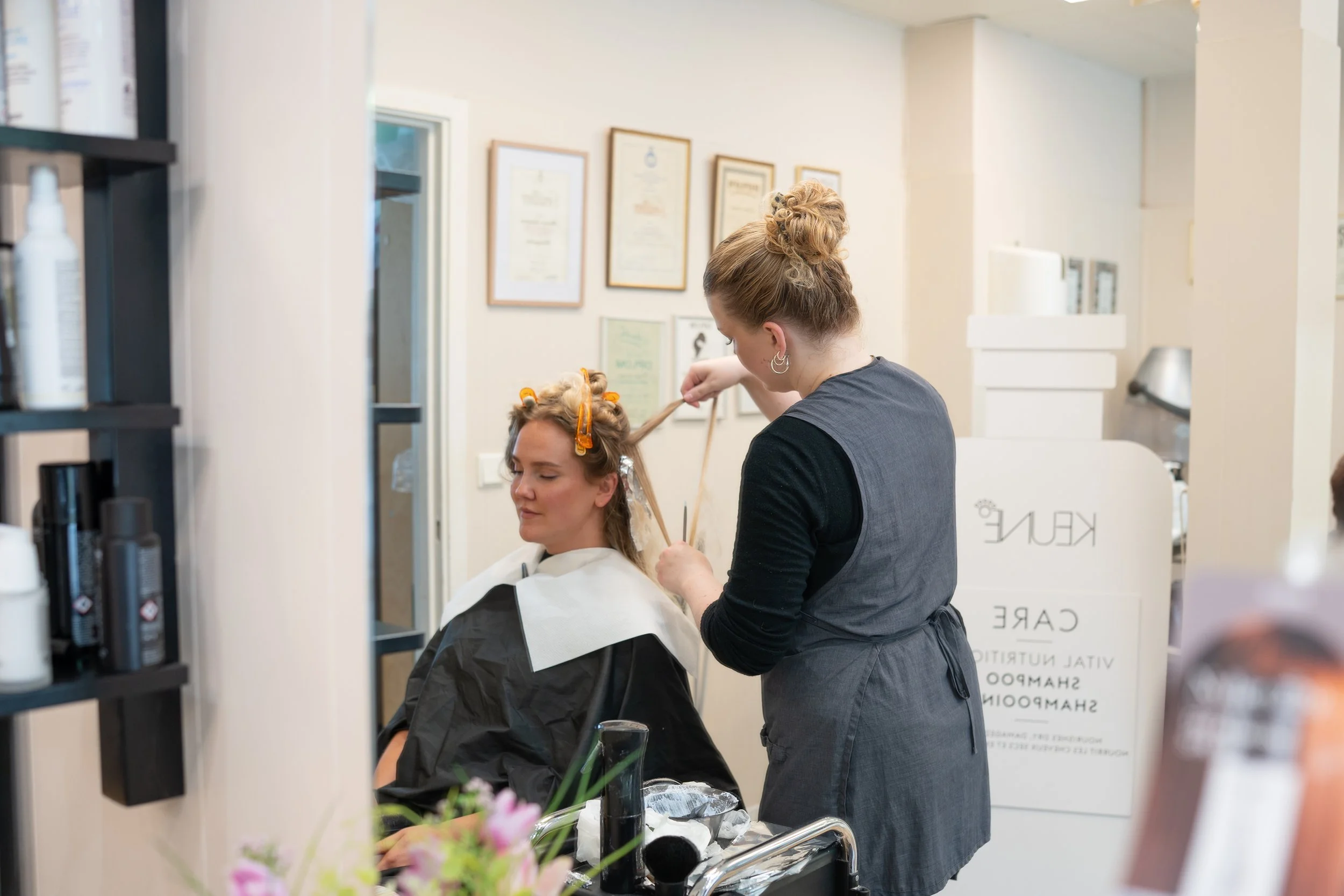 A woman receiving a hair treatment at a salon while a stylist applies hair color with a brush. The woman has hair rollers, and the stylist is in a gray apron. The salon has framed certificates on the wall and shelves with hair products.