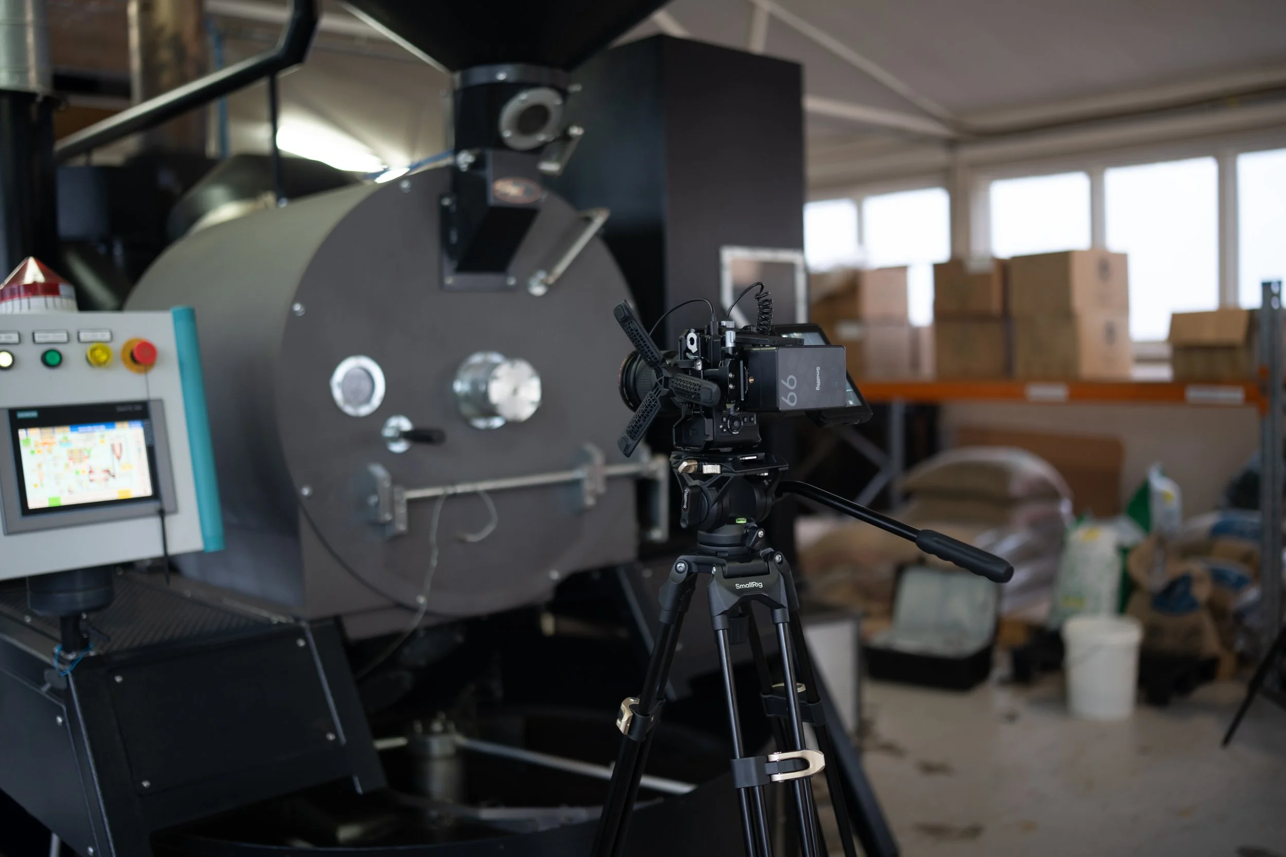 A professional camera on a tripod recording a large industrial machine in a warehouse with boxes and supplies in the background.