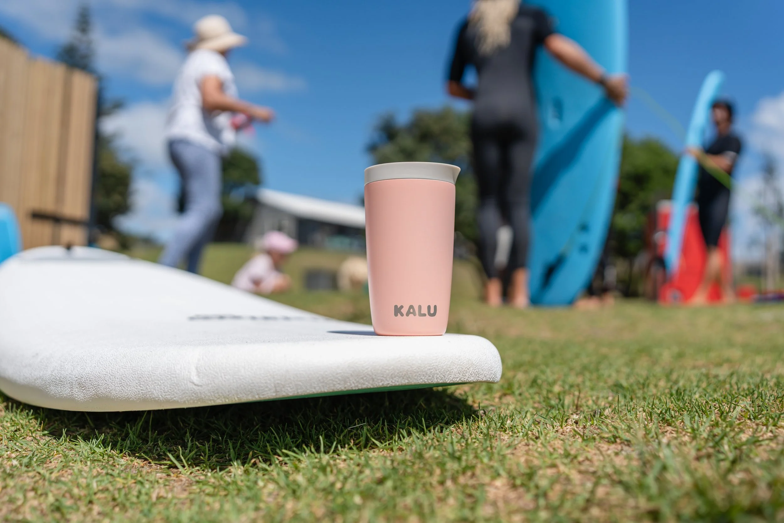 A pink KALU tumbler on a white surfboard on grass, with people holding surfboards and a woman in a black wetsuit preparing to surf in the background.
