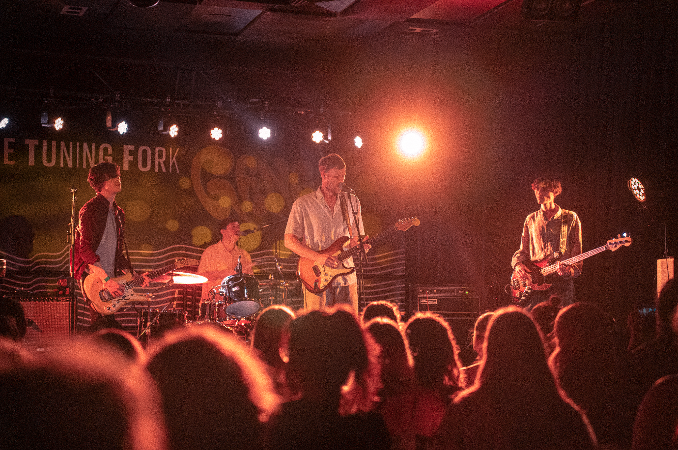 Four band members performing on stage during a live concert with audience in the foreground and stage lighting overhead.