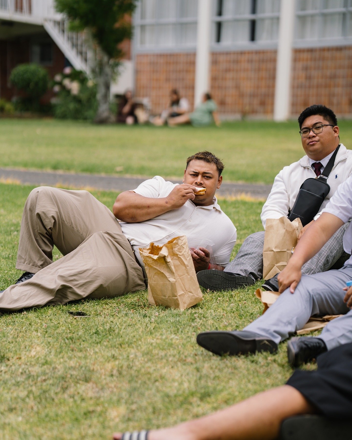 Group of young adults sitting and lying on grass during a picnic, with some eating from paper bags and others relaxing.