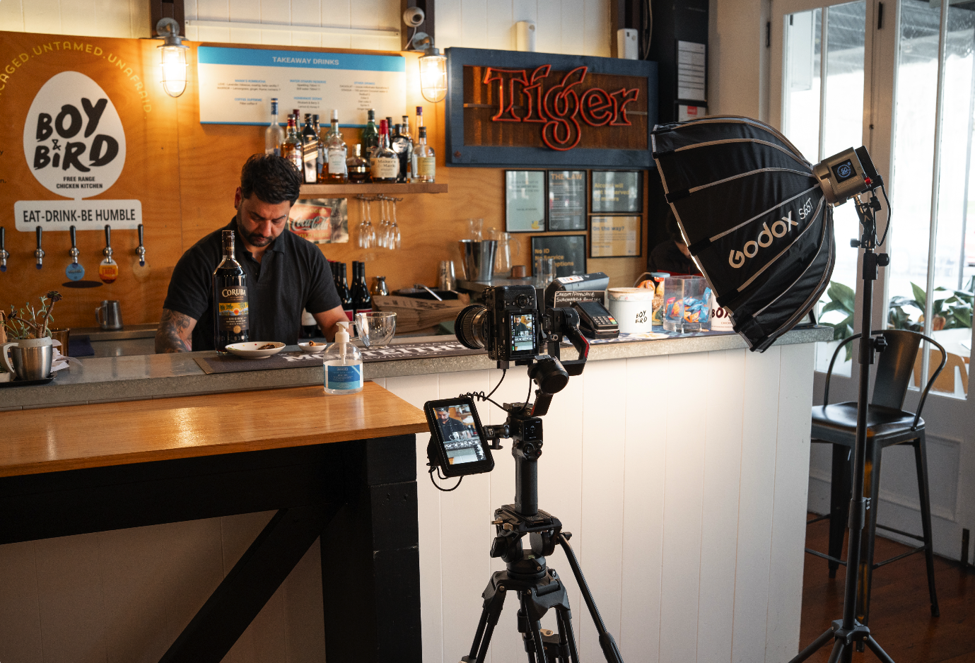 A man working behind a bar counter with camera and lighting equipment set up for filming or photography, inside a cafe or restaurant.