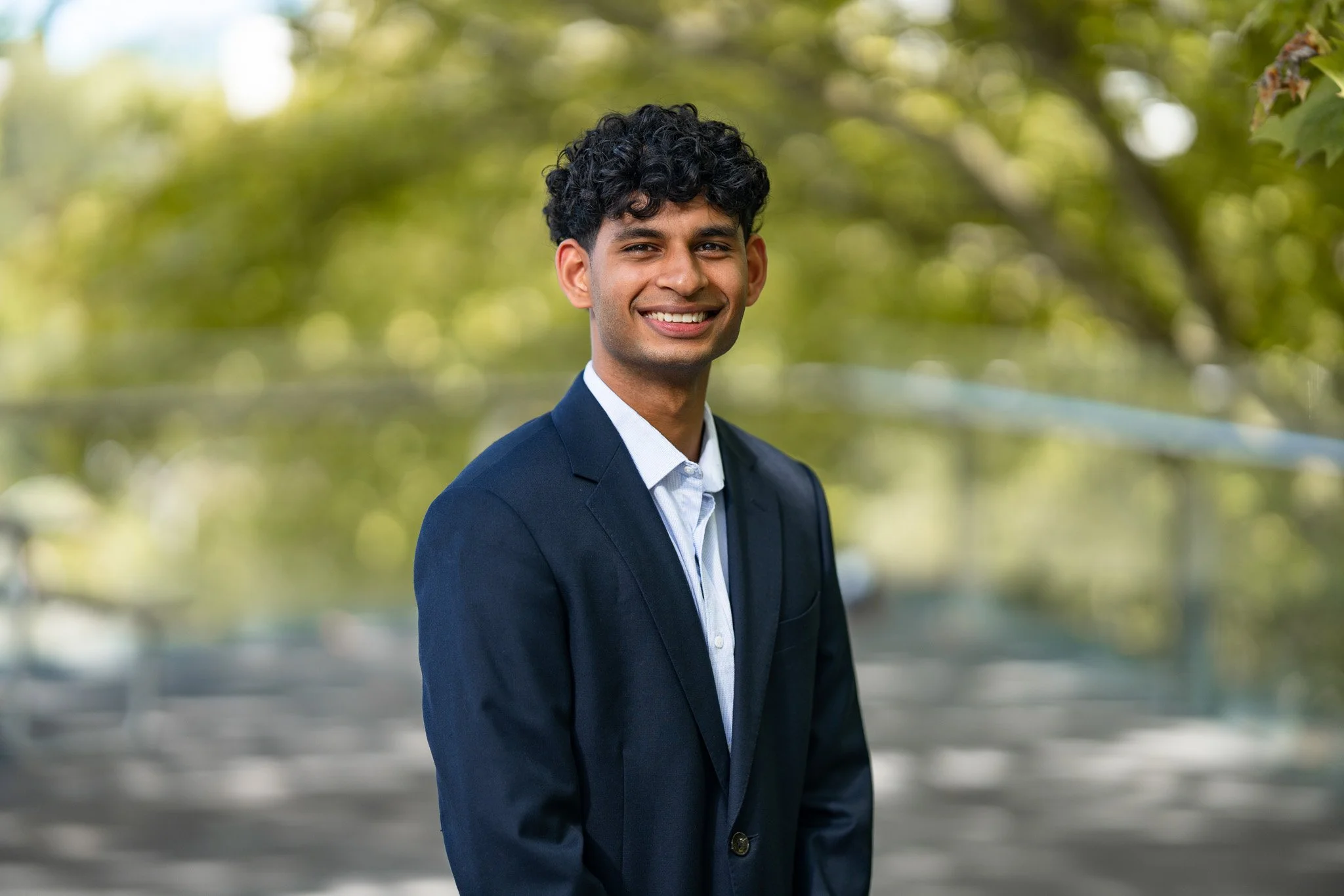 Young man in formal suit smiling outdoors with blurred green trees and sunlight in the background.