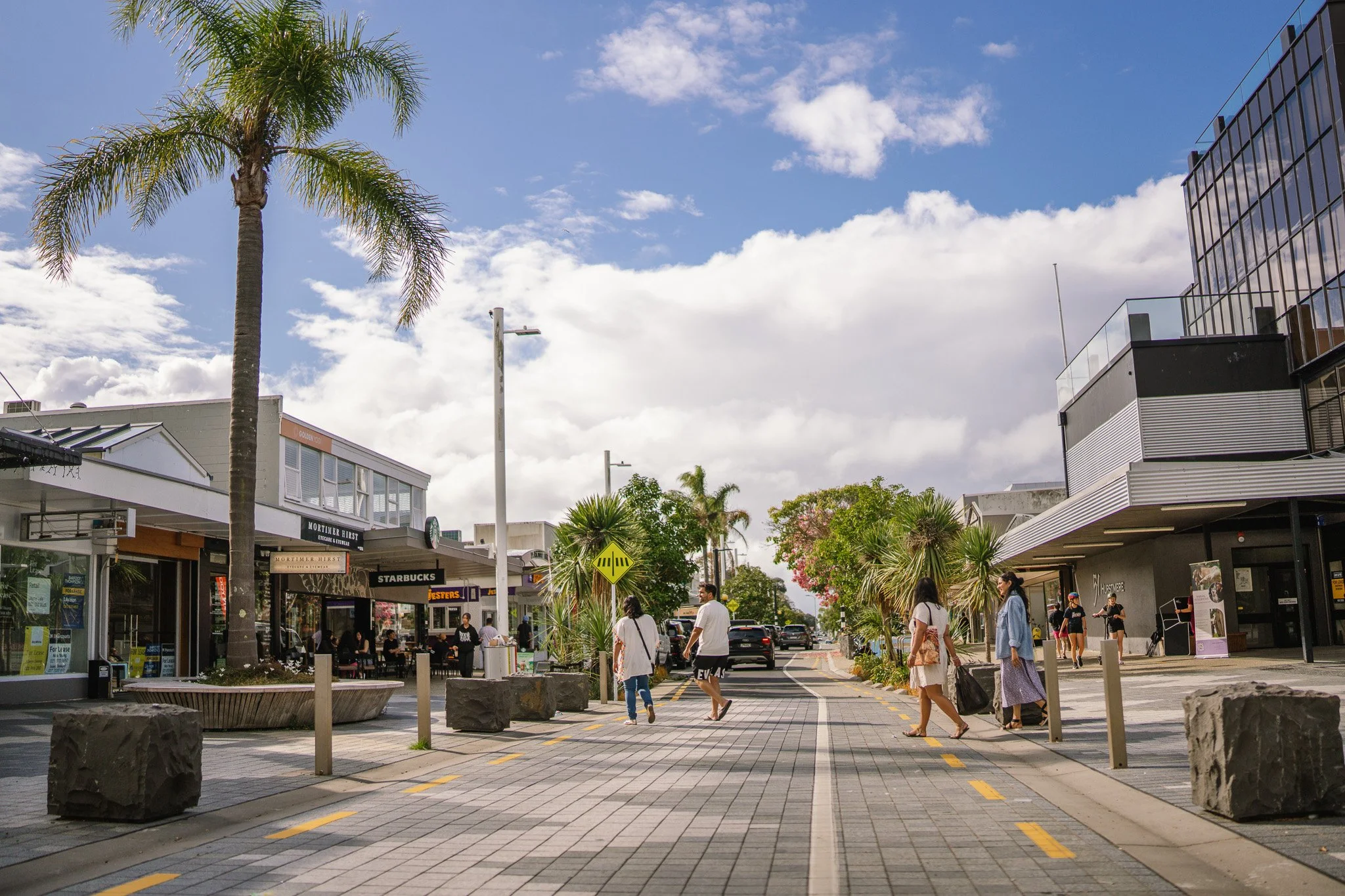 Shopping street with palm trees, shops, and people walking outdoors under a partly cloudy sky.