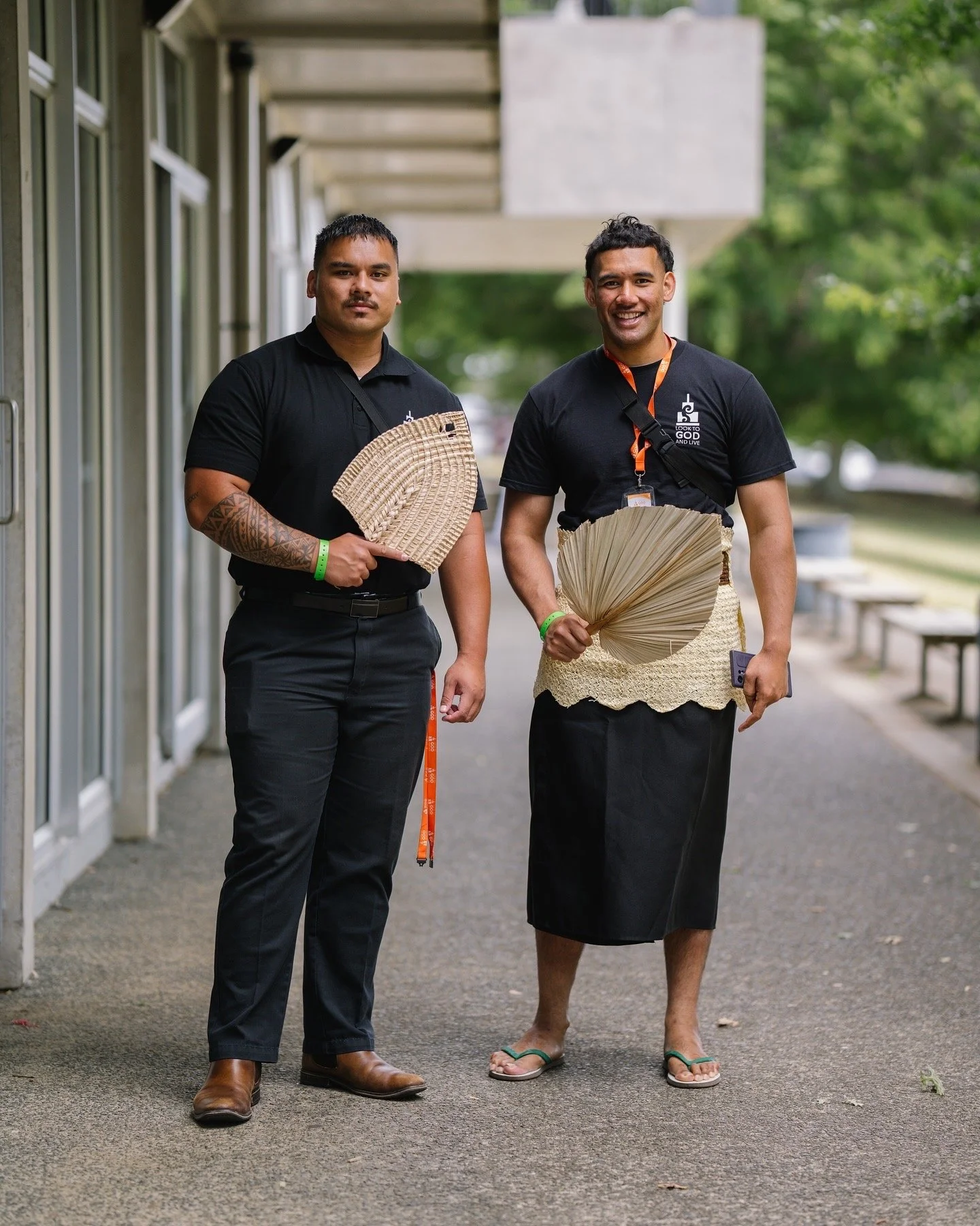 Two men standing outdoors on a sidewalk, holding woven fans. They are wearing black shirts, one with a skirt and the other with pants, and are smiling. Green trees are in the background.