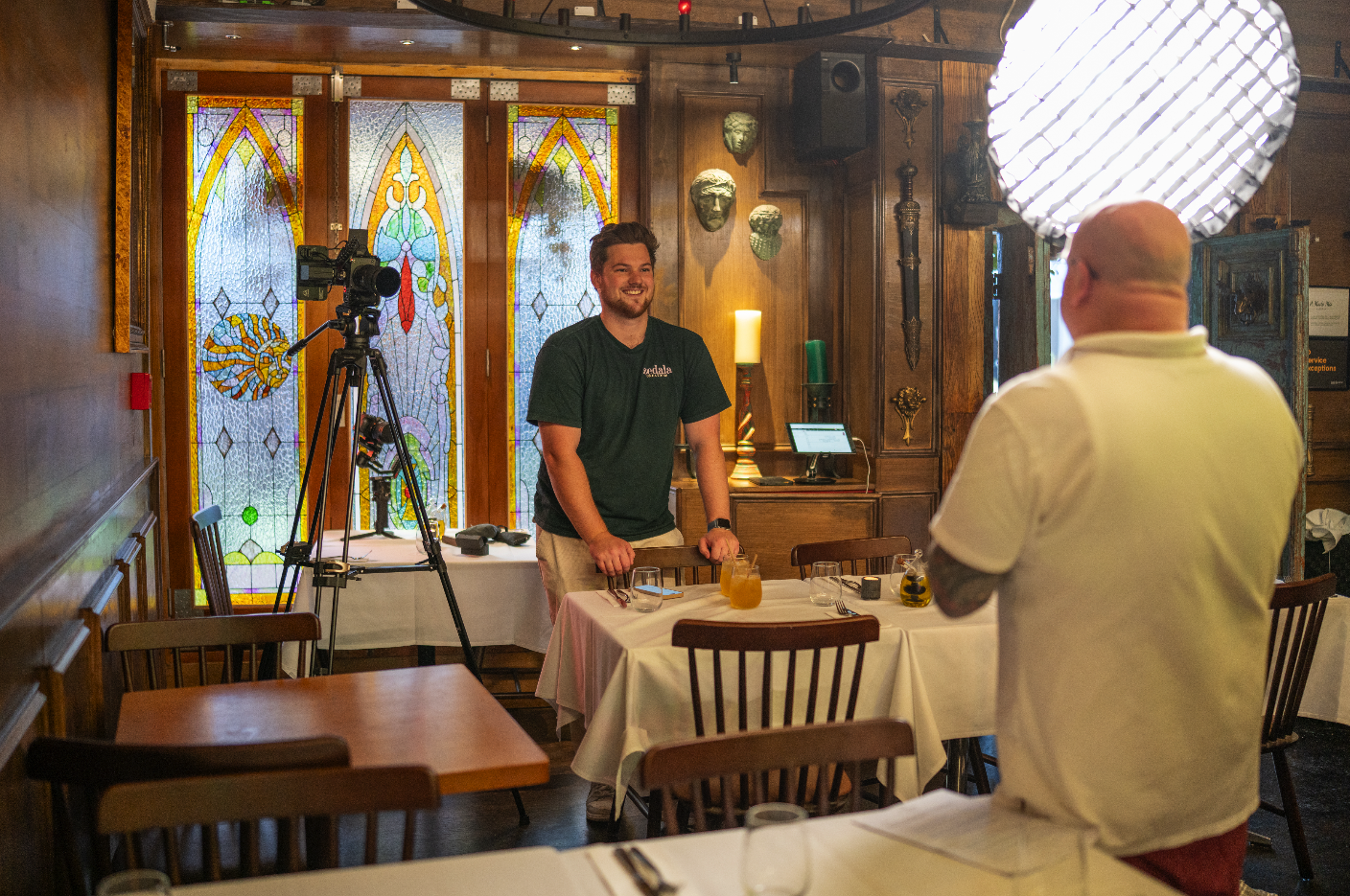 A man being interviewed by a person with a bald head in a restaurant decorated with stained glass windows and wooden accents.
