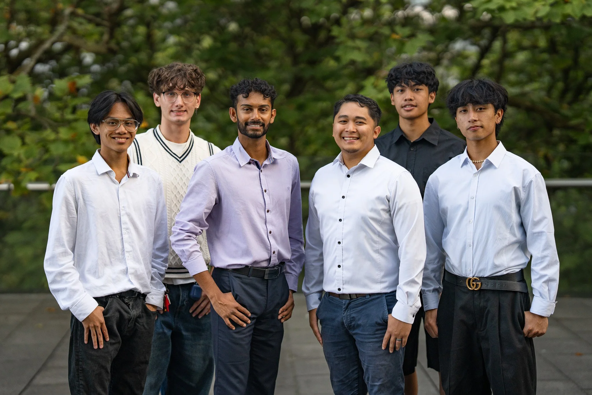 Six young men standing outdoors in front of green trees, dressed in semi-formal attire, smiling at the camera.